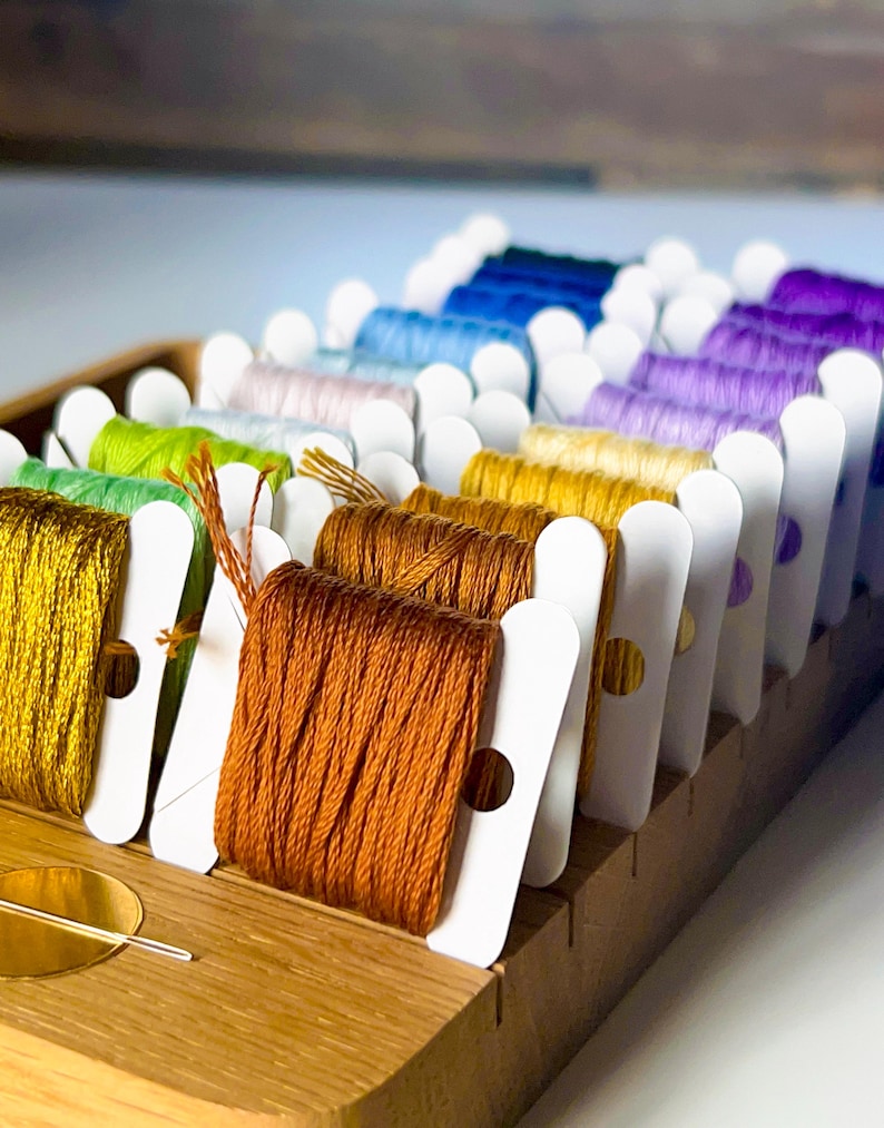 May include: A wooden organizer filled with spools of colorful embroidery floss. The floss is arranged in a rainbow of colors, including yellow, orange, green, blue, and purple. Each spool is neatly wound and held in place by a white card.