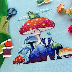 May include: A cross-stitch embroidery featuring a badger under a colorful mushroom with a frog on top. The mushroom is red, orange, and yellow with white spots. The badger is blue and purple with white stripes. The background is light blue with white rain drops.