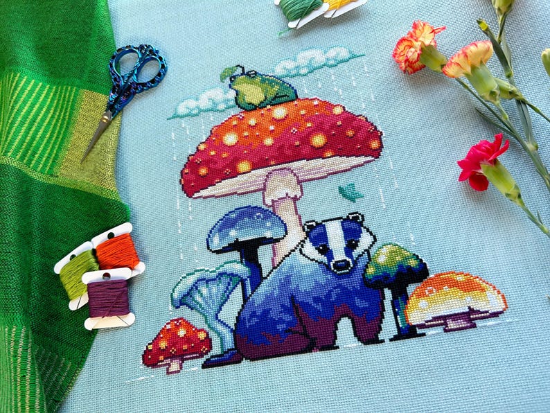 May include: A cross-stitch embroidery featuring a badger under a colorful mushroom with a frog on top. The mushroom is red, orange, and yellow with white spots. The badger is blue and purple with white stripes. The background is light blue with white rain drops.