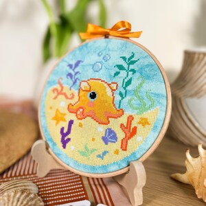 May include: A yellow and orange octopus with a blue background, surrounded by coral, seaweed, and bubbles. The octopus is embroidered on a round canvas with a wooden frame.