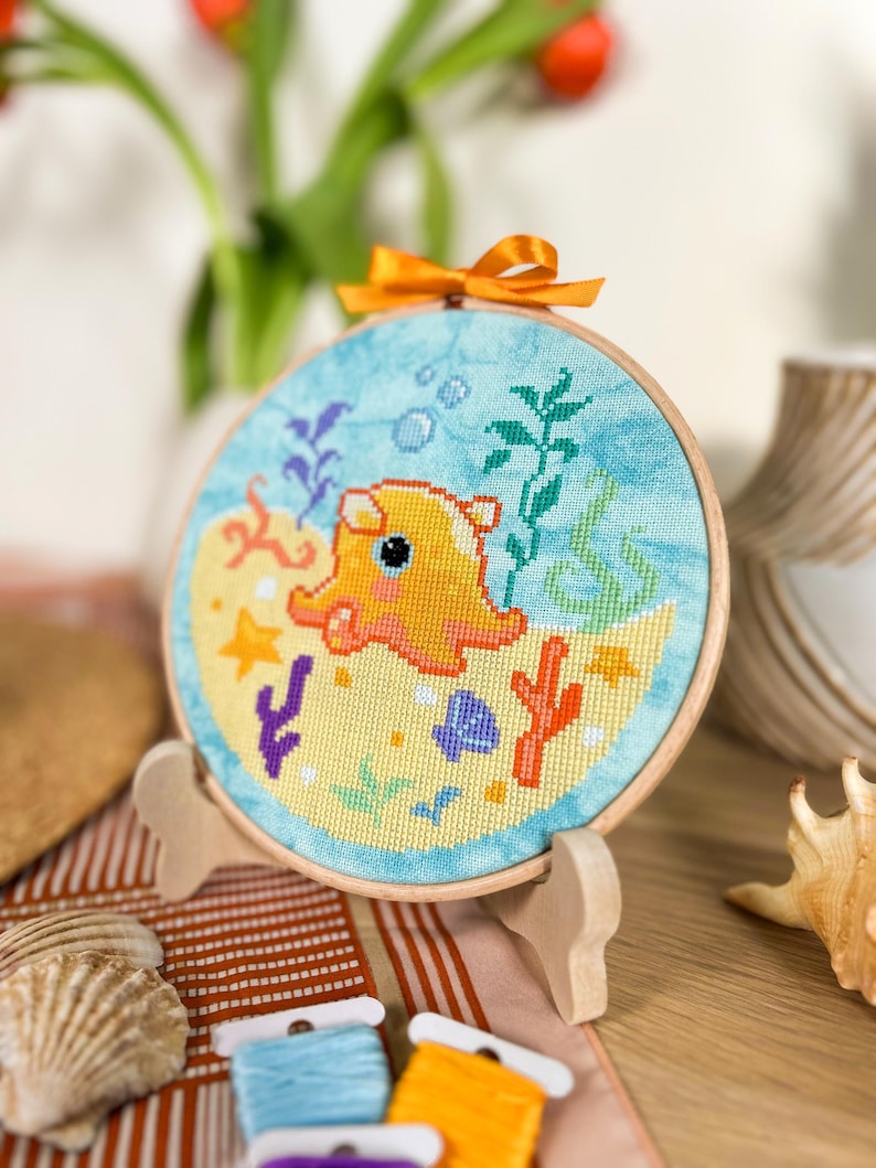 May include: A yellow and orange octopus with a blue background, surrounded by coral, seaweed, and bubbles. The octopus is embroidered on a round canvas with a wooden frame.