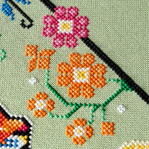 May include: Close-up of a cross-stitch floral design on a light green fabric. The design features pink, orange, and white flowers with green stems and leaves. A black diagonal line and blue accents are also present.