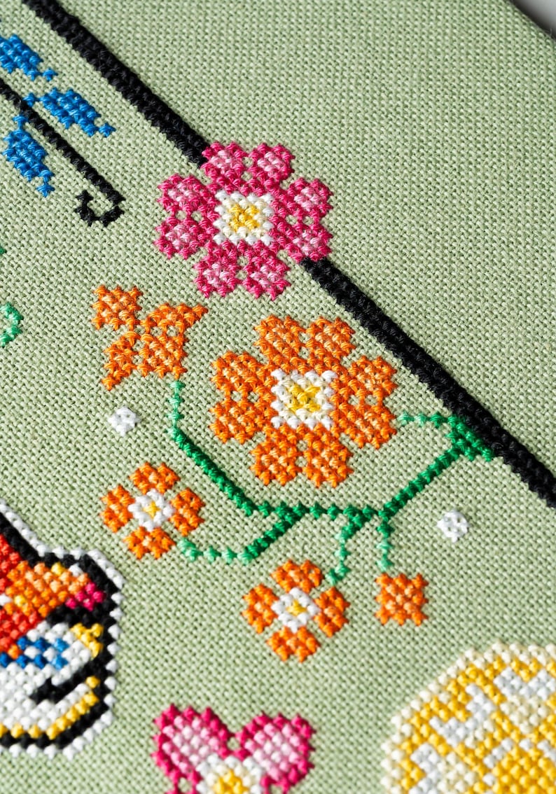 May include: Close-up of a cross-stitch floral design on a light green fabric. The design features pink, orange, and white flowers with green stems and leaves. A black diagonal line and blue accents are also present.