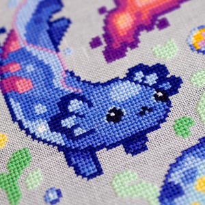 May include: Close-up of a cross-stitch design featuring a blue axolotl with pink accents. The design includes other pixelated elements in shades of purple, pink, yellow, and green on a gray fabric background.