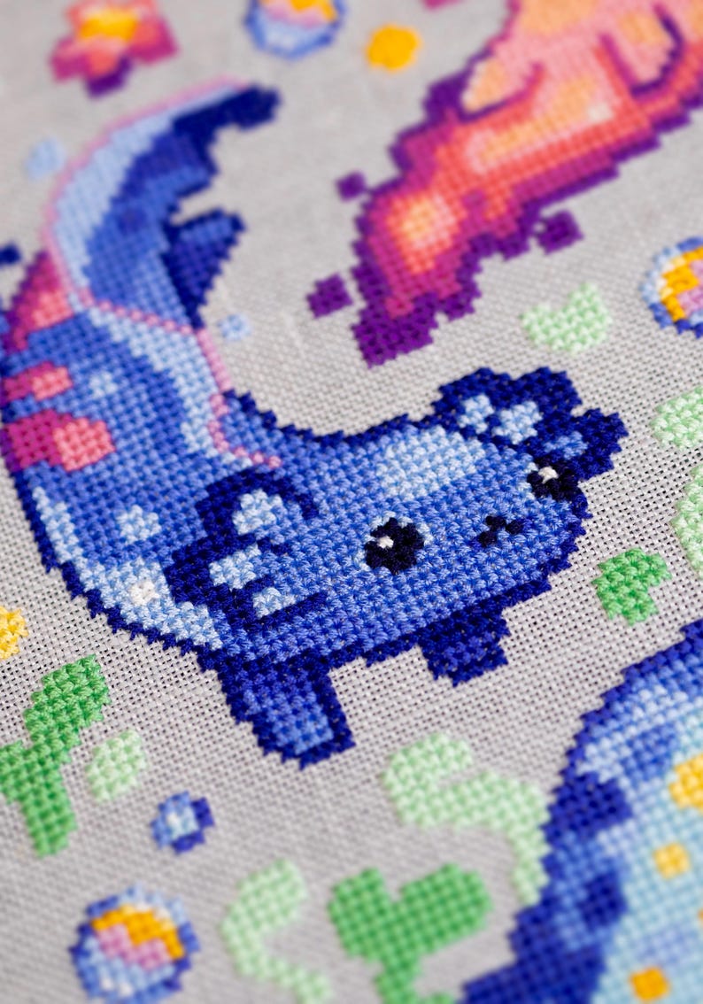 May include: Close-up of a cross-stitch design featuring a blue axolotl with pink accents. The design includes other pixelated elements in shades of purple, pink, yellow, and green on a gray fabric background.