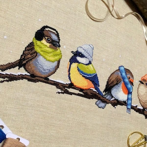 Closeup of Winter Birds cross stitch pattern. Image is zoomed in on the sparrow. He is a very cute bird that also looks very hip. The bird is wearing a green scarf and earmuffs, and looks very warm. The colors of his feathers are brown, beige, grey.