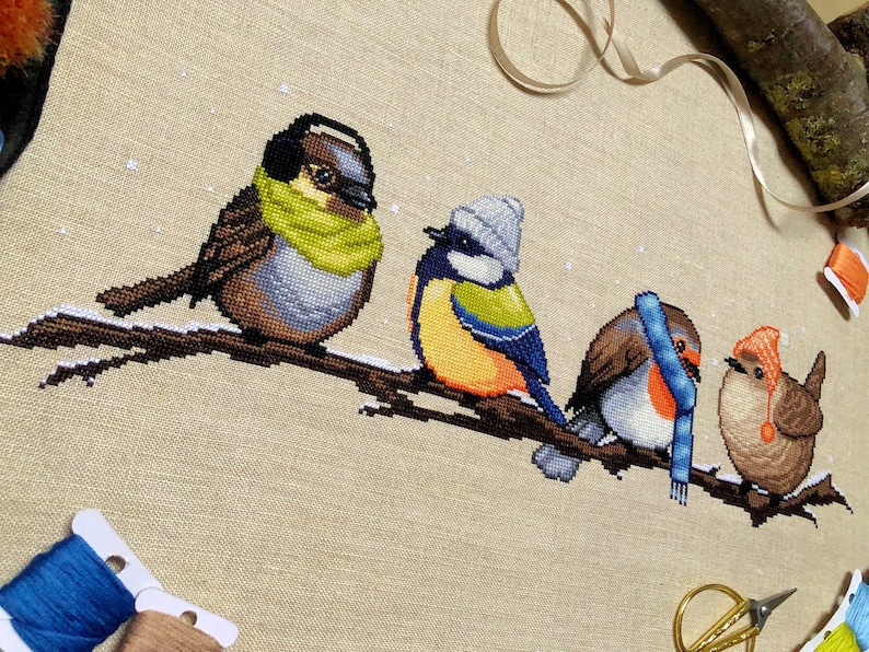 Closeup of Winter Birds cross stitch pattern. Image is zoomed in on the sparrow. He is a very cute bird that also looks very hip. The bird is wearing a green scarf and earmuffs, and looks very warm. The colors of his feathers are brown, beige, grey.