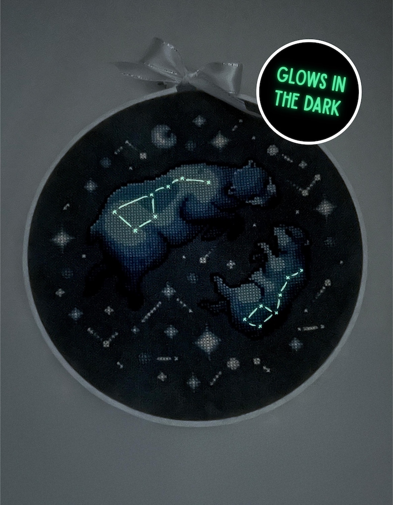 Puede incluir: Un bordado de punto de cruz redondo, azul oscuro, con un dise&ntilde;o de oso y constelaciones. El bordado brilla en la oscuridad, con el texto "GLOWS IN THE DARK" en un c&iacute;rculo. Una cinta blanca est&aacute; atada en la parte superior.