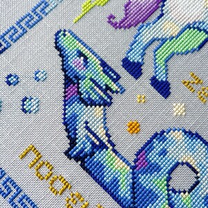May include: A cross-stitch design featuring a blue and purple dragon with green and yellow accents. The design includes the word "nostalgia" in yellow and a blue border with Greek key patterns. The background is a light gray fabric.