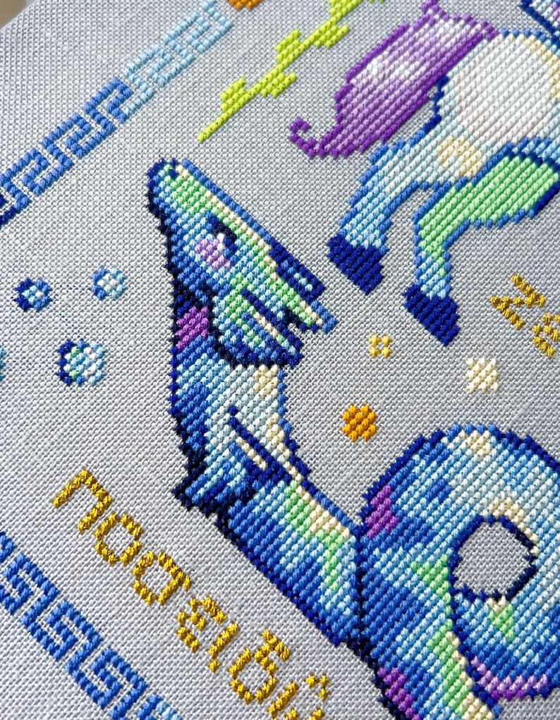 May include: A cross-stitch design featuring a blue and purple dragon with green and yellow accents. The design includes the word "nostalgia" in yellow and a blue border with Greek key patterns. The background is a light gray fabric.