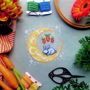Front view of Dreaming of Carrots cross stitch pattern. Scene is surrounded by flowers, carrots and embroidery supplies. Moon is round but is surrounded by white stars that create a square composition. Bunny looks peaceful and content.