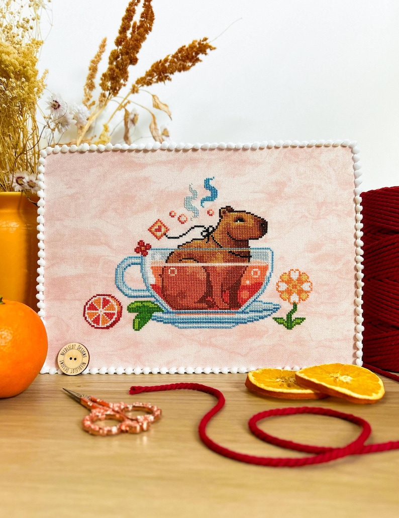 K&ouml;nnte beinhalten: Kreuzstich-Stickerei mit einem Capybara in einer Teetasse vor einem rosa Hintergrund. Das Capybara ist braun und hat einen wei&szlig;en Streifen um den Hals. Die Teetasse ist mit einer roten Fl&uuml;ssigkeit gef&uuml;llt und es steigt Dampf aus ihr auf. Auf dem Tisch liegen auch eine kleine Orangenscheibe und eine Blume.