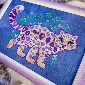 Puede incluir: Arte de punto de cruz de un leopardo estilizado en tonos morados y beige, sobre un fondo de tela azul. La pieza enmarcada presenta un marco de color lavanda y detalles florales verdes. Una artesan&iacute;a &uacute;nica hecha a mano.