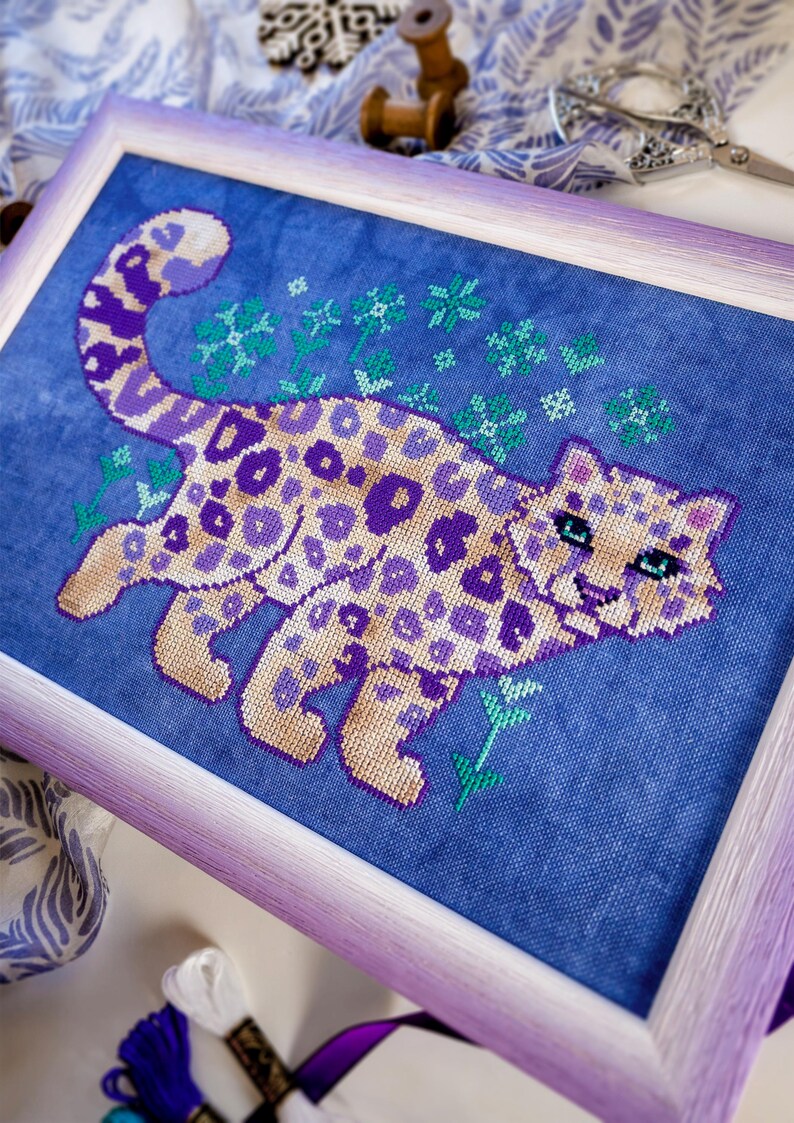 Puede incluir: Arte de punto de cruz de un leopardo estilizado en tonos morados y beige, sobre un fondo de tela azul. La pieza enmarcada presenta un marco de color lavanda y detalles florales verdes. Una artesan&iacute;a &uacute;nica hecha a mano.