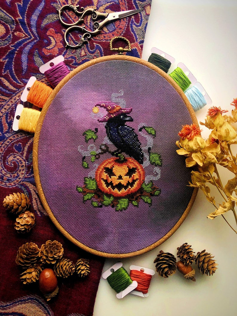 以下が含まれることがあります： ハロウィーンのデザインが施されたクロスステッチ刺繍枠。魔女の帽子をかぶった黒いカラスが彫刻されたカボチャの上に止まっているデザインです。背景は紫色のグラデーション。刺繍糸とハサミが枠を囲んでいます。