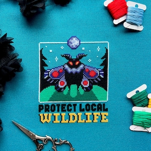 May include: A cross-stitch design on turquoise fabric featuring a black moth with red and purple accents, set against a green landscape and a starry sky. The text "PROTECT LOCAL WILDLIFE" is stitched below. Spools of thread and scissors are also visible.