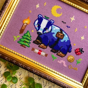 Puede incluir: Arte de punto de cruz de un tej&oacute;n azul y blanco con una mochila, rodeado de una antorcha, un &aacute;rbol, una luna y estrellas. La obra de arte est&aacute; enmarcada en un marco dorado y marr&oacute;n ornamentado, sobre un fondo lavanda.