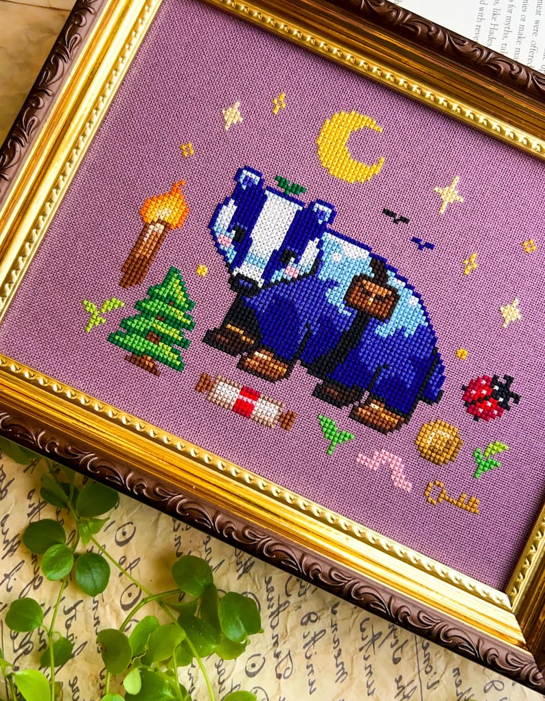 Puede incluir: Arte de punto de cruz de un tej&oacute;n azul y blanco con una mochila, rodeado de una antorcha, un &aacute;rbol, una luna y estrellas. La obra de arte est&aacute; enmarcada en un marco dorado y marr&oacute;n ornamentado, sobre un fondo lavanda.