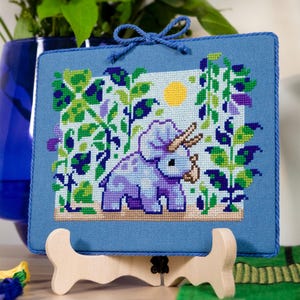 Puede incluir: Un bordado de punto de cruz con un triceratops azul claro en una escena de jard&iacute;n con un sol amarillo. La obra de arte est&aacute; enmarcada en tela azul claro y se exhibe en un caballete de madera. La escena incluye follaje verde y morado.