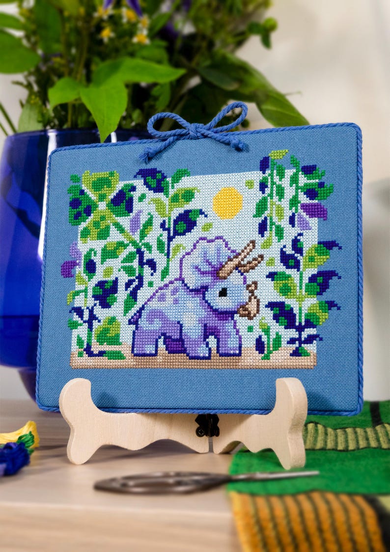 Puede incluir: Un bordado de punto de cruz con un triceratops azul claro en una escena de jard&iacute;n con un sol amarillo. La obra de arte est&aacute; enmarcada en tela azul claro y se exhibe en un caballete de madera. La escena incluye follaje verde y morado.