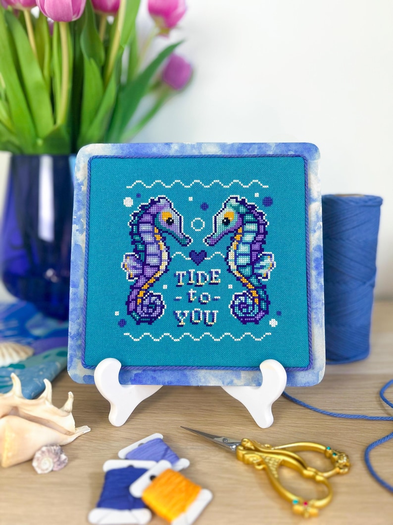 May include: Cross-stitch art featuring two seahorses facing each other, with the words "Tide to You" stitched below. The seahorses are purple and blue on a turquoise background, framed in a blue marbled border. The artwork is displayed on a white easel.