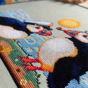 May include: Cross-stitch embroidery of two puffins with colorful beaks and a yellow sun in the background. The puffins are perched on a green and brown rock with blue and white waves behind them.