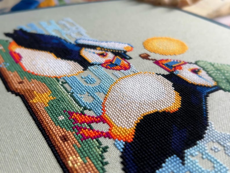 May include: Cross-stitch embroidery of two puffins with colorful beaks and a yellow sun in the background. The puffins are perched on a green and brown rock with blue and white waves behind them.