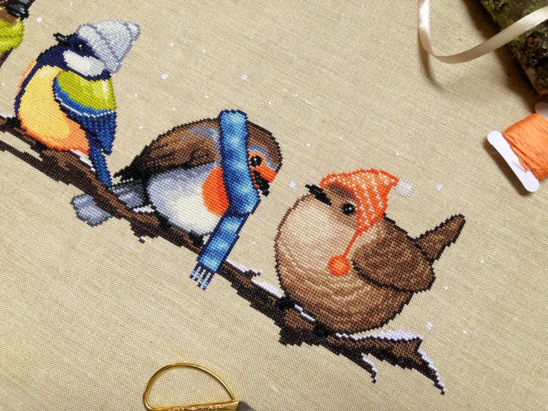 Closeup of Winter Birds cross stitch pattern. Two of the four birds are depicted, these are the robin and wren. Colors are yellow, brown, orange, blue, green and white. Robin is wearing a scarf and the wren is wearing a hat with dangly pompoms.