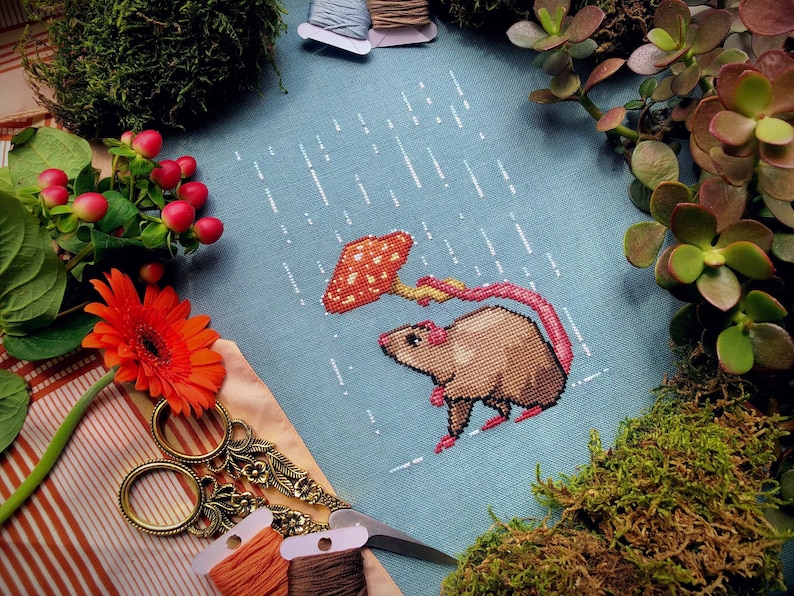 May include: A cross-stitch pattern of a brown mouse holding a red and orange mushroom umbrella in a blue rain-themed background.