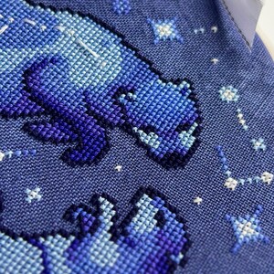 Puede incluir: Primer plano de un dise&ntilde;o de punto de cruz con un oso en tonos azules y morados sobre un fondo azul oscuro. Estrellas blancas y constelaciones est&aacute;n dispersas alrededor del oso. El dise&ntilde;o est&aacute; sobre una tela texturizada.