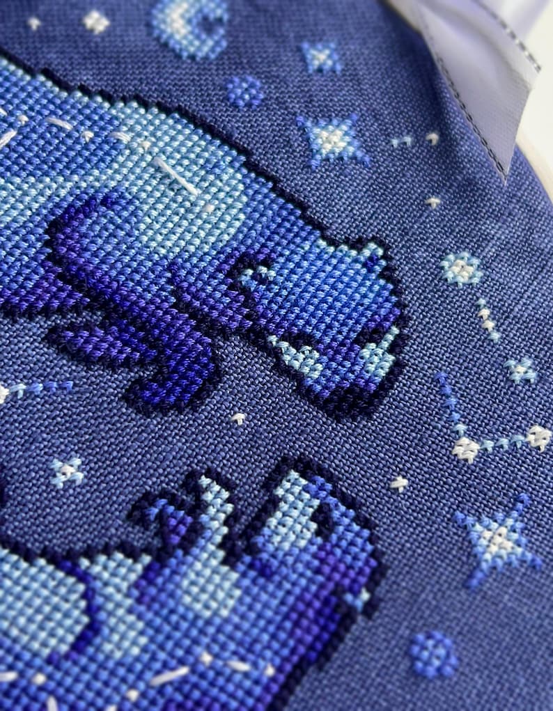 Puede incluir: Primer plano de un dise&ntilde;o de punto de cruz con un oso en tonos azules y morados sobre un fondo azul oscuro. Estrellas blancas y constelaciones est&aacute;n dispersas alrededor del oso. El dise&ntilde;o est&aacute; sobre una tela texturizada.