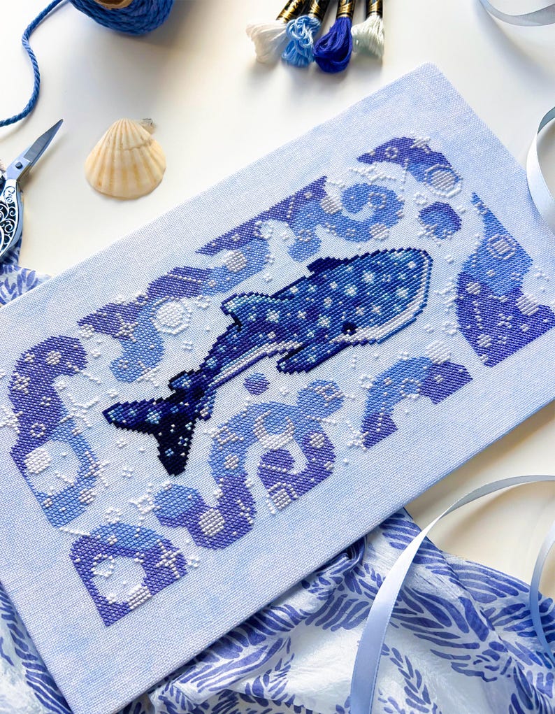 May include: Cross-stitch art featuring a whale shark in shades of blue and white. The design includes a detailed whale shark with a dotted pattern, surrounded by stylized waves. The piece is on light blue fabric, with scissors, thread, and a seashell nearby.