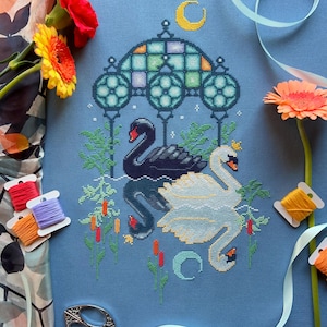 Puede incluir: Bordado de punto de cruz con dos cisnes, uno negro y otro blanco, bajo un arco de vitral con una luna creciente arriba. El dise&ntilde;o est&aacute; en una tela azul con acentos verdes, amarillos y rojos.