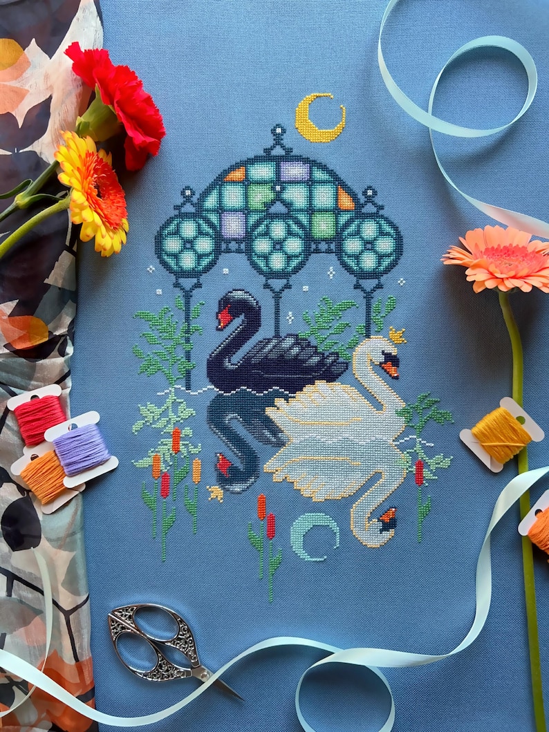 Puede incluir: Bordado de punto de cruz con dos cisnes, uno negro y otro blanco, bajo un arco de vitral con una luna creciente arriba. El dise&ntilde;o est&aacute; en una tela azul con acentos verdes, amarillos y rojos.