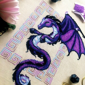 Flat-lay of Dragon cross stitch pattern. Stitched item, surrounded by decorations. Finished piece is of medium to large size. Colors are blue, purple, white, black and pink. Dragon is surrounded by variegated frame. Its head is like that of a horse.