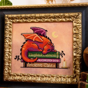 May include: Cross-stitch art of a red and purple dragon resting on a stack of books with titles like "Flight Manual" and "Alchemy 101." The framed artwork has a gold ornate frame and a candle is lit nearby. The background is a peach color.
