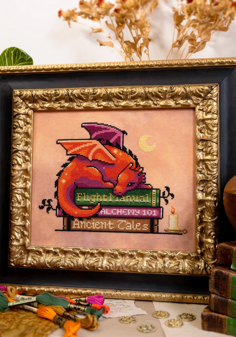 May include: Cross-stitch art of a red and purple dragon resting on a stack of books with titles like "Flight Manual" and "Alchemy 101." The framed artwork has a gold ornate frame and a candle is lit nearby. The background is a peach color.