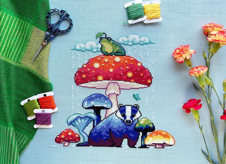 May include: Cross-stitch embroidery of a badger sitting under a red and white mushroom with a green frog on top. The mushroom is surrounded by smaller mushrooms in shades of blue and yellow. The background is a light blue fabric with a rain pattern.