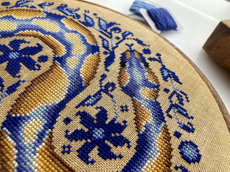 Peut inclure: Gros plan d'un projet de broderie au point de croix termin&eacute; repr&eacute;sentant un motif de serpent stylis&eacute; dans des tons de bleu, jaune et marron. La broderie est encadr&eacute;e dans un cercle en bois.