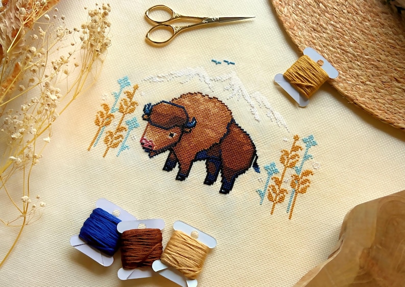 May include: Cross-stitch embroidery of a brown bison standing in front of a snowy mountain range with yellow and blue wildflowers. The embroidery is on a cream-colored fabric.