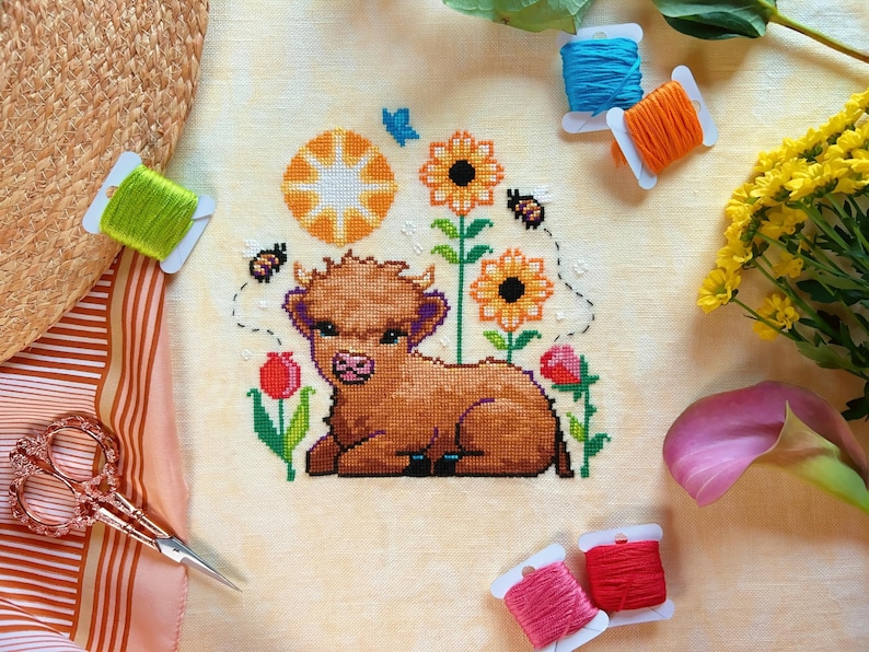 May include: A cross-stitch embroidery of a brown Highland cow with a yellow sun and sunflowers in the background. The embroidery is surrounded by colorful embroidery floss.