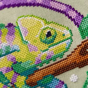 May include: Close-up of a cross-stitch design featuring a chameleon. The chameleon is primarily yellow and green, with purple accents. It is perched on a brown branch, with a light green background. The stitching is detailed and colorful.