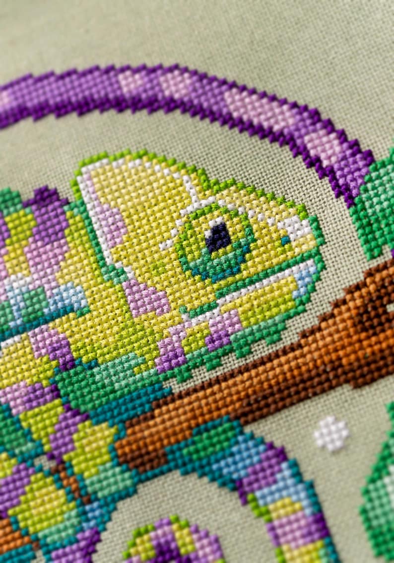 May include: Close-up of a cross-stitch design featuring a chameleon. The chameleon is primarily yellow and green, with purple accents. It is perched on a brown branch, with a light green background. The stitching is detailed and colorful.