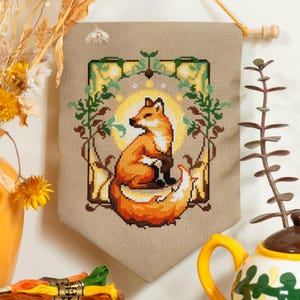 May include: A cross-stitch wall hanging featuring a fox. The fox is orange and white, set against a beige background with a yellow sun and green foliage. The hanging is suspended by a wooden dowel and string.