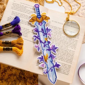 May include: A handmade, cross-stitched bookmark in the shape of a sword, featuring a blue blade with purple floral accents and a gold hilt. The bookmark rests on an open book with colorful embroidery floss nearby.