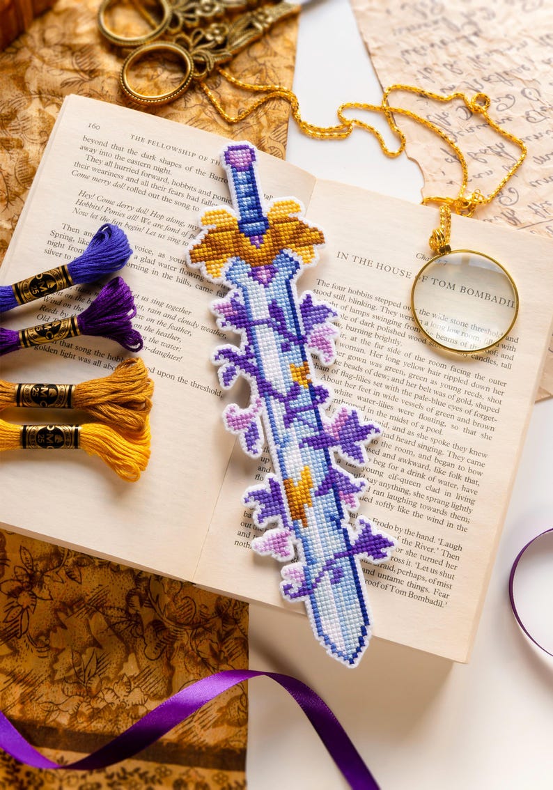 Puede incluir: Marcap&aacute;ginas artesanal en forma de espada, bordado a punto de cruz, con una hoja azul, motivos florales morados y una empu&ntilde;adura dorada. Descansa sobre un libro abierto, con hilo de bordar colorido cerca.