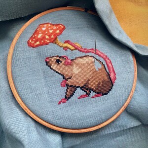 Image of the rat with mushroom umbrella being stitched. Stitches are neat and tidy. Fabric is placed inside an embroidery hoop. Colors are bright and beautiful: pink, brown, red, orange and yellow. Rat is looking cute and happy.