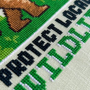 May include: Close-up of a cross-stitch design with the words "PROTECT LOCAL WILDLIFE" in black and green. The design features brown animal legs on a green background, stitched on a cream-colored fabric.