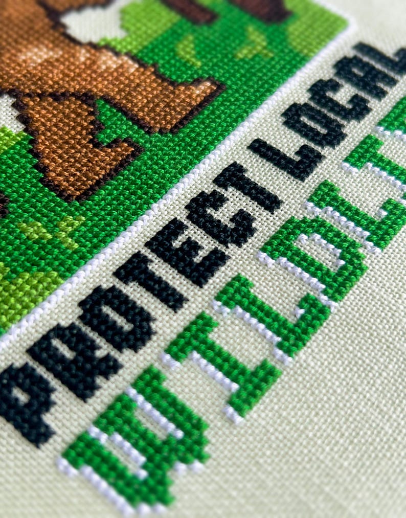May include: Close-up of a cross-stitch design with the words "PROTECT LOCAL WILDLIFE" in black and green. The design features brown animal legs on a green background, stitched on a cream-colored fabric.