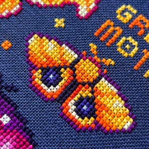 Op de afbeelding: Close-up van een kruissteekontwerp met een kleurrijke vlinder met oranje, gele en paarse tinten op een donkerblauwe achtergrond. De tekst "GR MOT" is in oranje gestikt. Het ontwerp bevat sterren en andere geometrische vormen.